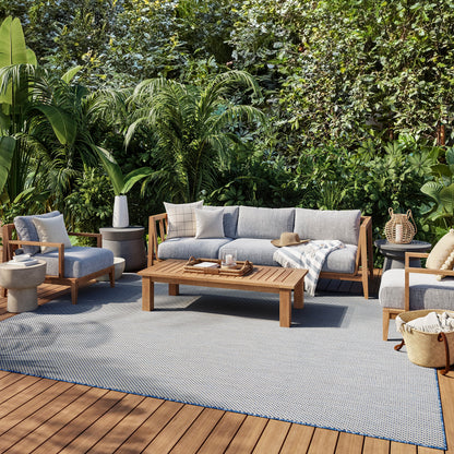 Outdoor lounge set with gray cushions and wooden frames, coffee table on a blue-gray rug amid tropical plants.