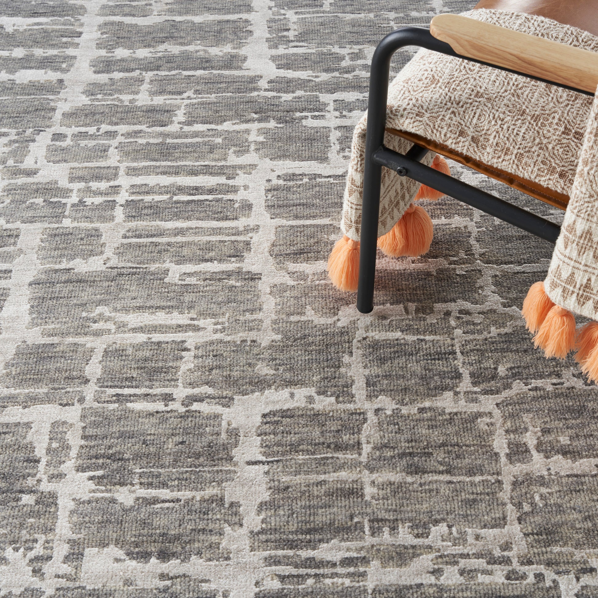 Gray woven rug with a beige geometric grid pattern under a chair with black metal legs and orange tassel fringe.