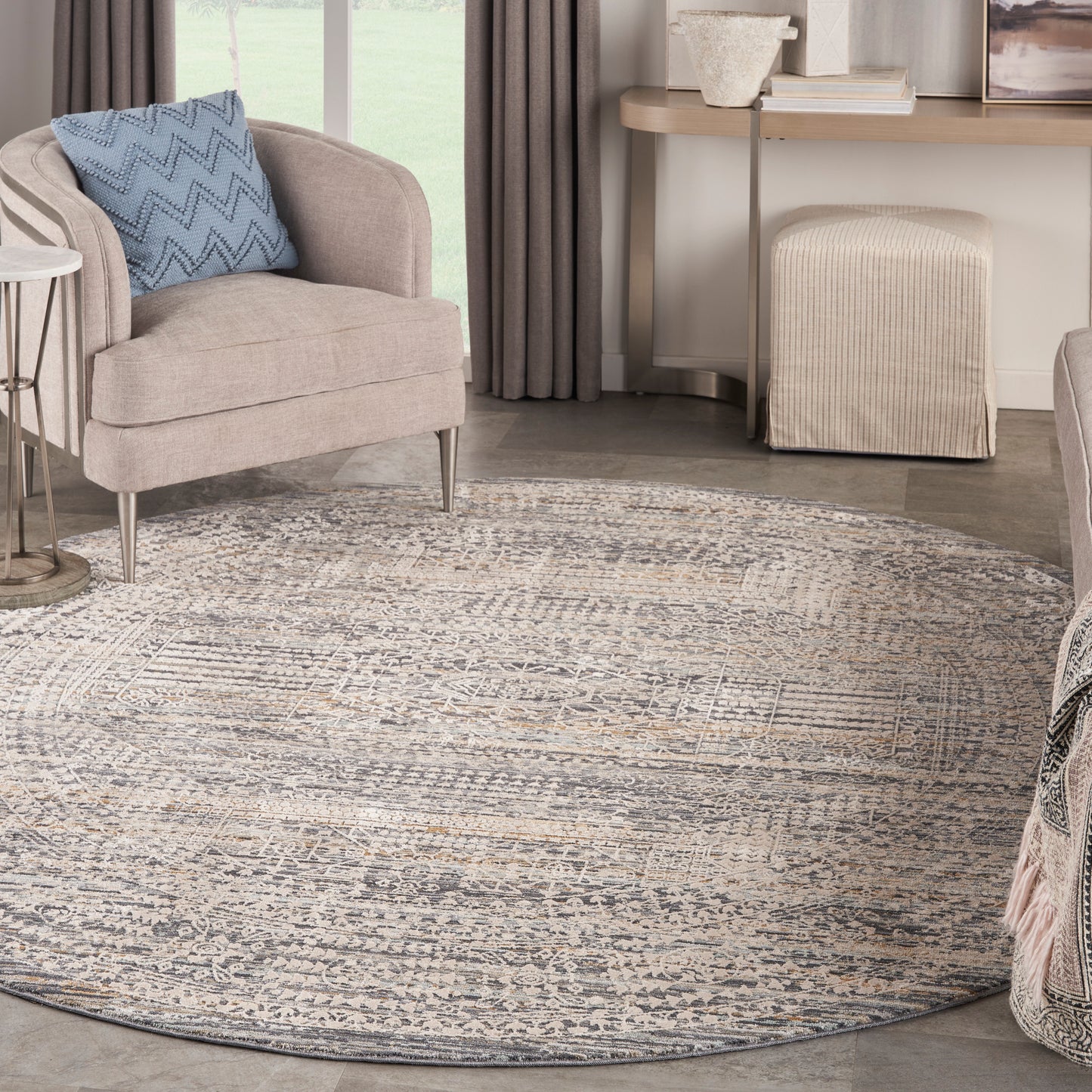 Round gray-beige patterned area rug under a beige upholstered armchair with a blue chevron pillow in a contemporary living room.