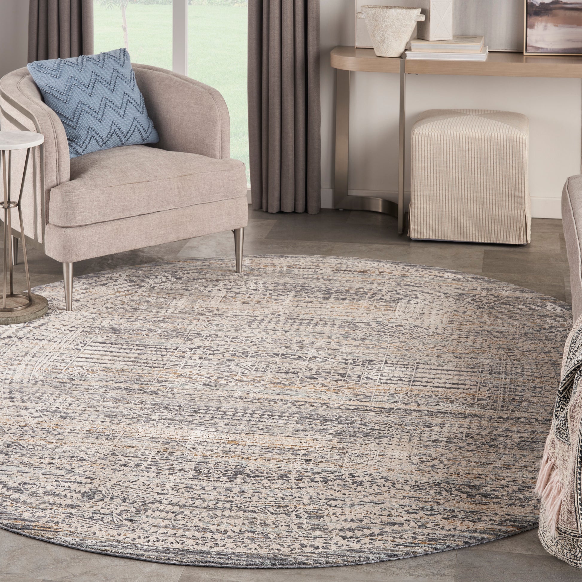 Round gray-beige patterned area rug under a beige upholstered armchair with a blue chevron pillow in a contemporary living room.