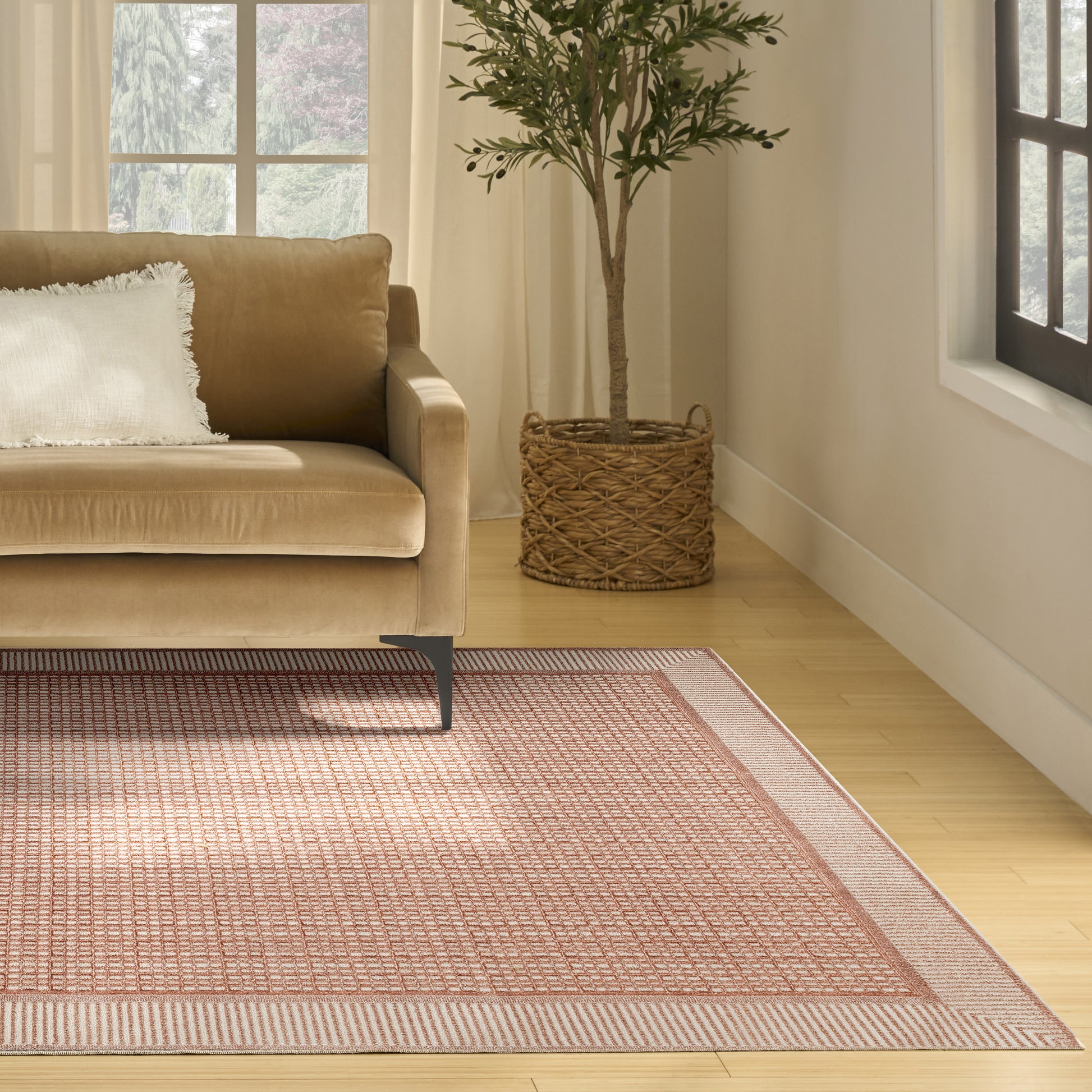 Beige sofa with white pillow in living room; tall plant in a wicker basket beside a pink-bordered rug on wood floor.