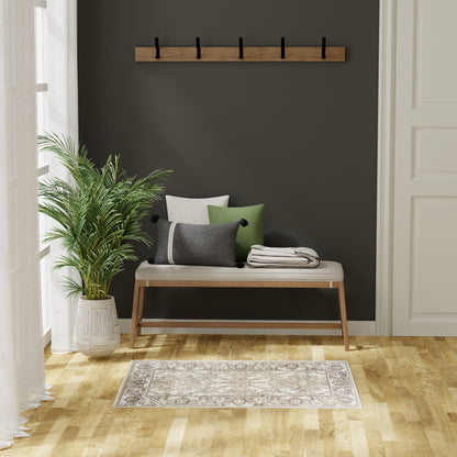 Wood-framed bench with light cushion and green/gray pillows against a dark wall, coat rack above, potted plant, beige patterned rug.