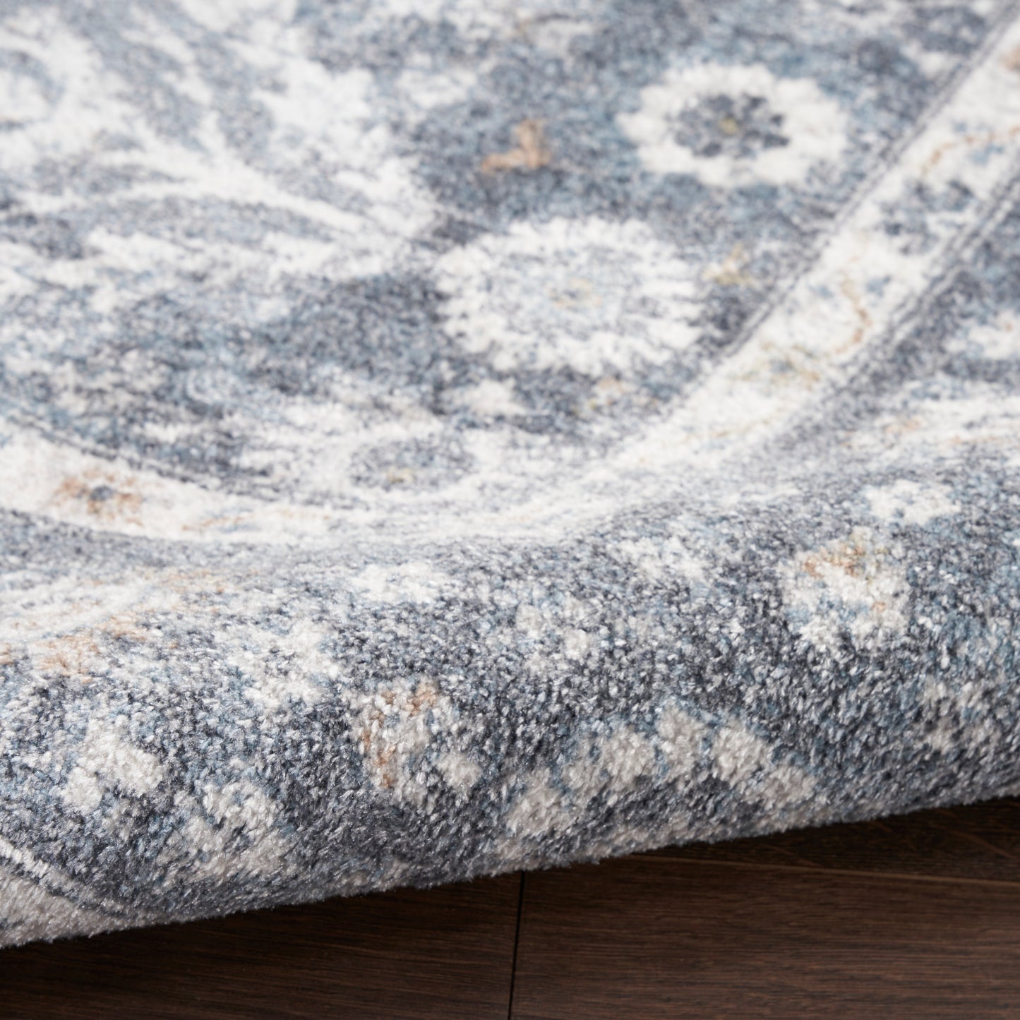 Soft blue and ivory patterned rug with a distressed medallion design, folded corner on dark wood floor.