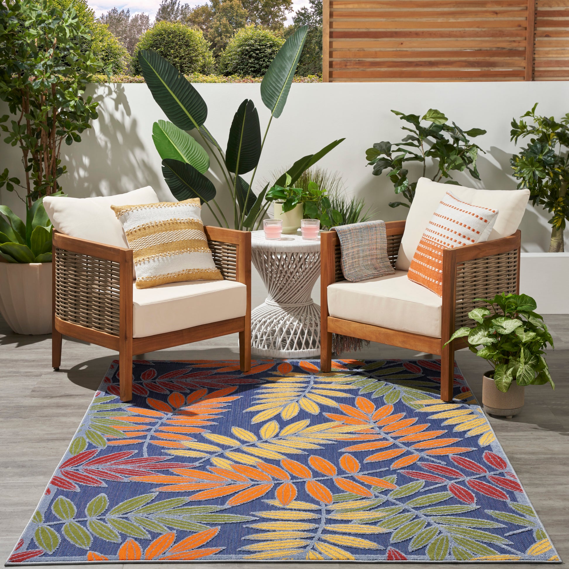 Two wood-frame outdoor chairs & white cushions & striped pillows, white woven table, bold tropical leaf rug amid plants.