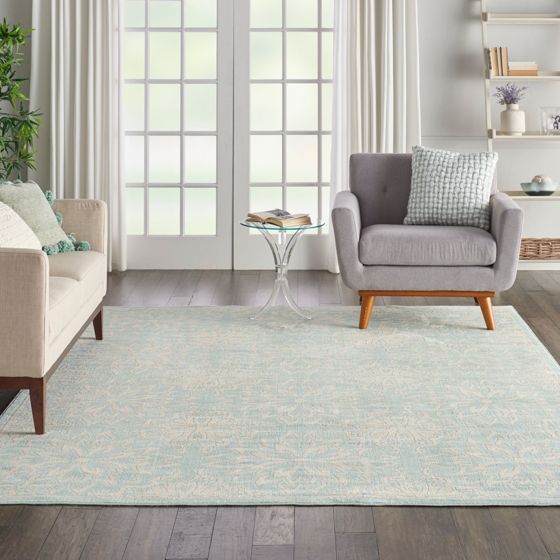 Bright living room with pale blue patterned rug, beige sofa, gray armchair with wooden legs, glass side table, curtains.