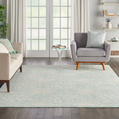 Bright living room with pale blue patterned rug, beige sofa, gray armchair with wooden legs, glass side table, curtains.