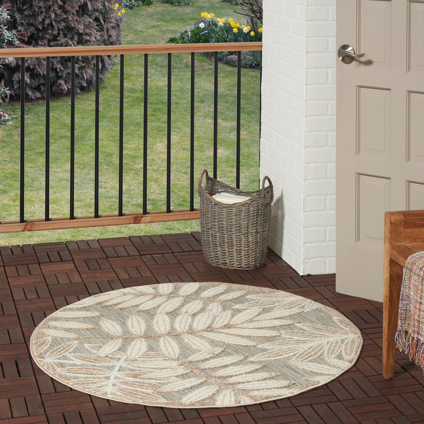 Round beige outdoor rug with a leaf pattern on a wooden deck beside a wicker basket and doorway.