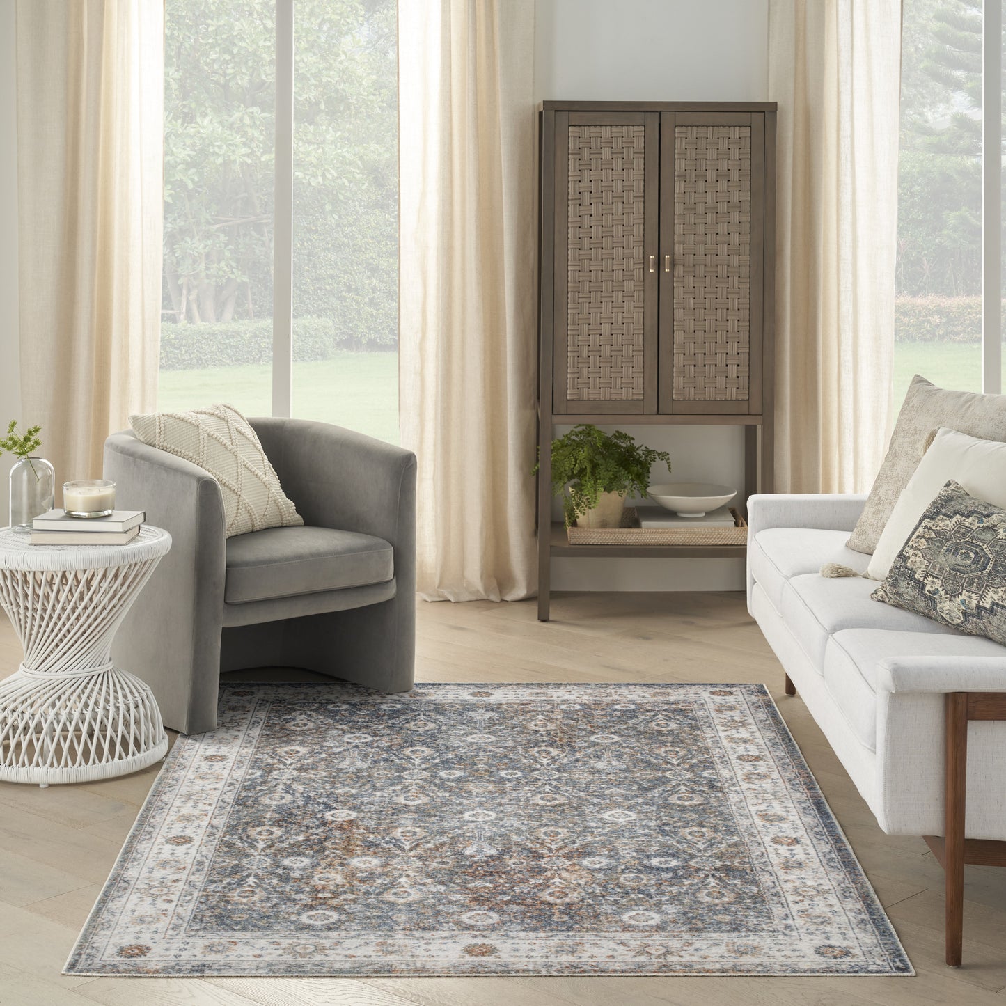 Bright living room with gray armchair, white sofa, woven cabinet, plant, and a blue-gray patterned area rug.