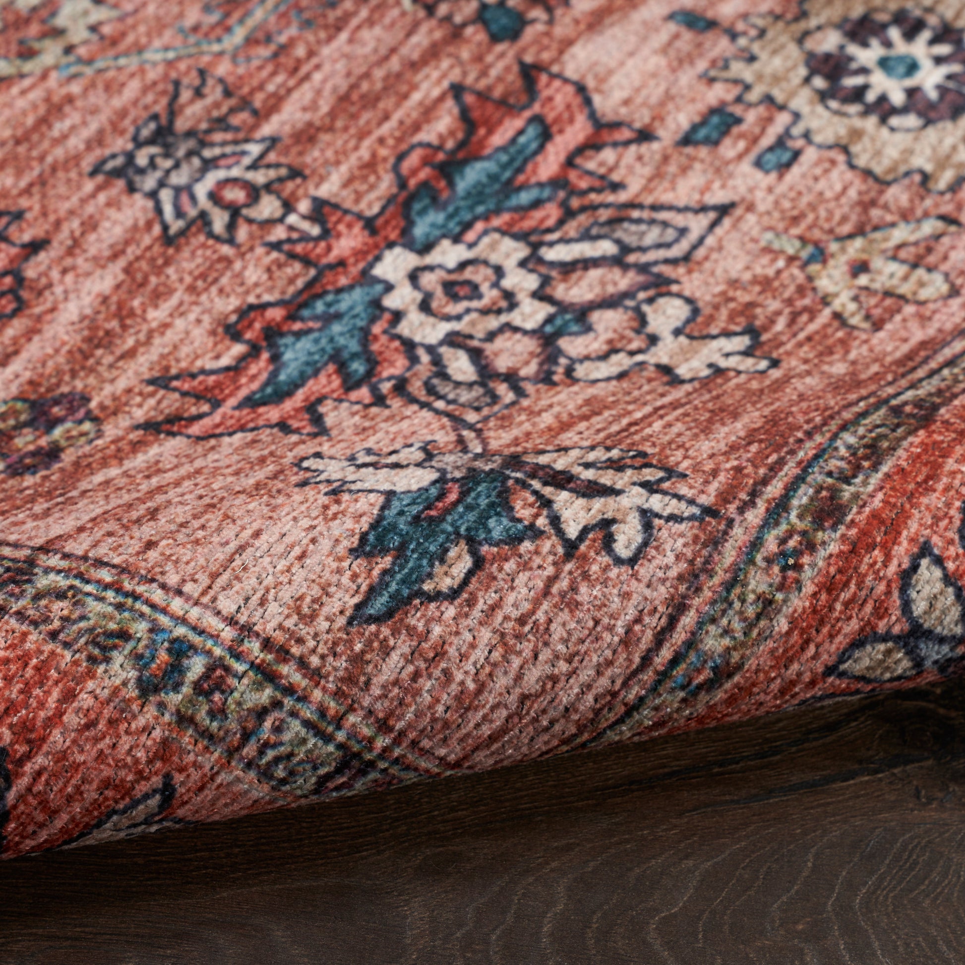 Close-up of rust-orange oriental rug with blue and cream floral motifs and a decorative border, folded on dark wood floor.