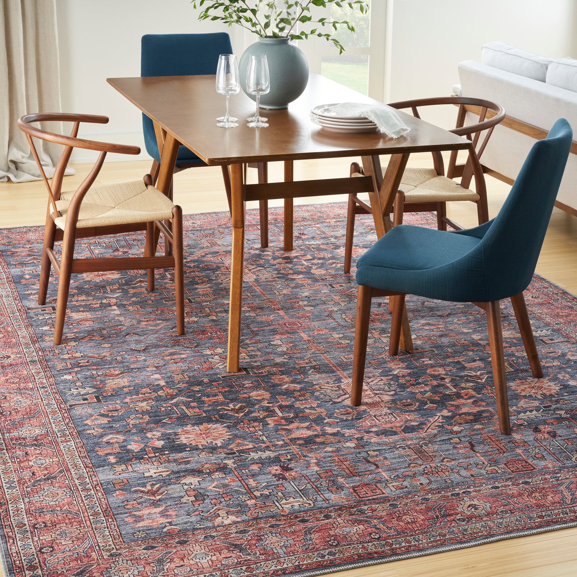 Mid-century wooden dining table with blue upholstered and cane-seat chairs on a red oriental rug.
