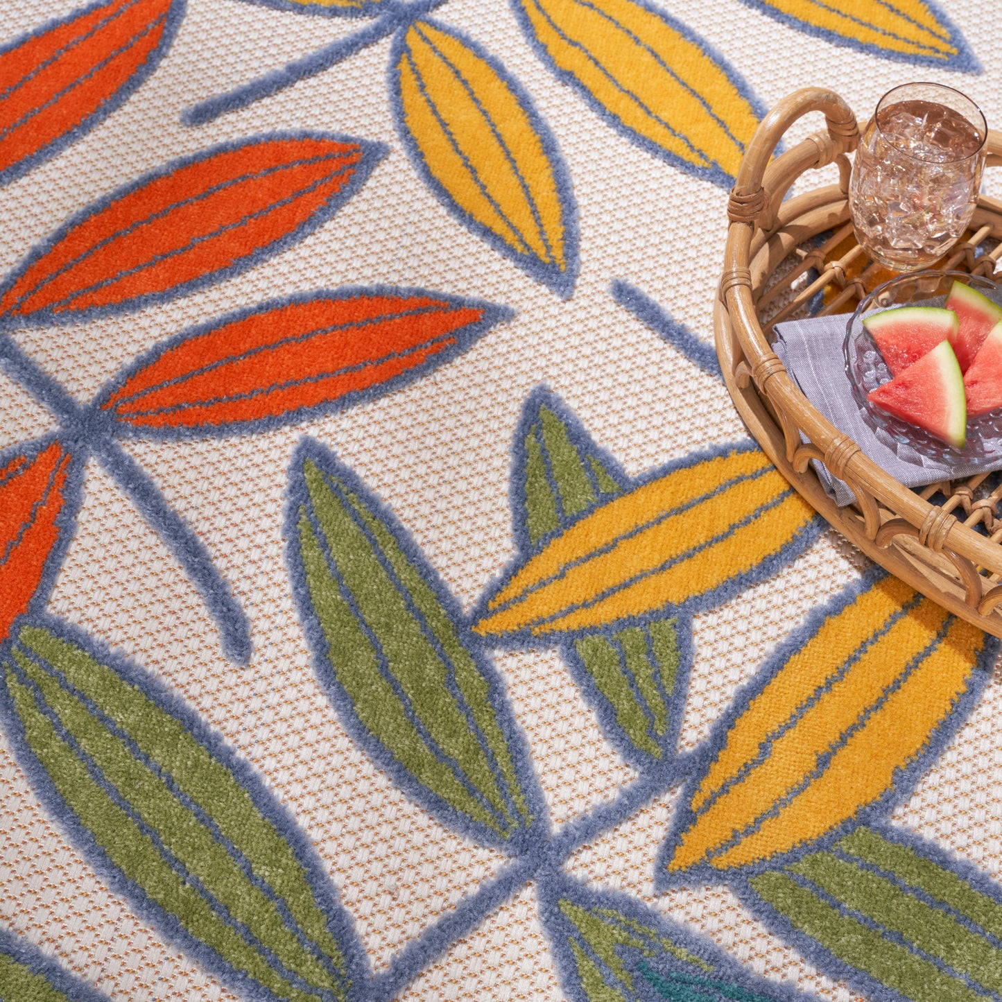 Close-up of a woven beige tropical rug with orange, yellow and green leaf motifs; a wicker basket and watermelon slices.