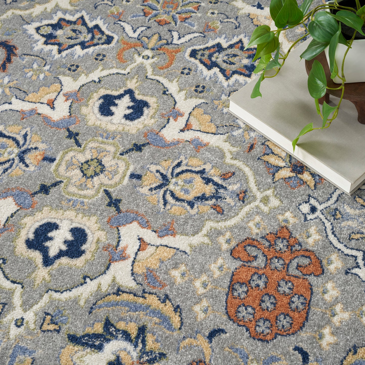 Intricate gray-blue floral patterned rug with cream, navy and tan accents; potted plant sits on beige books nearby.
