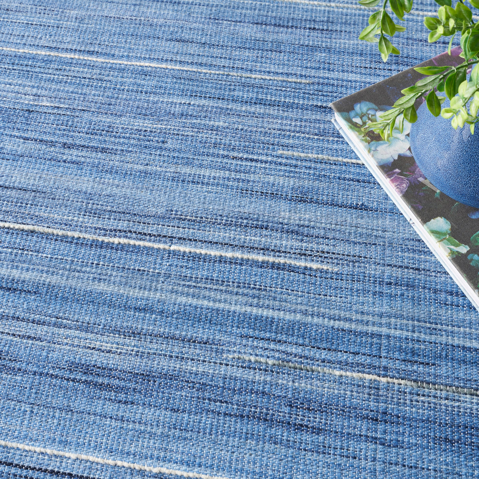 Blue woven striped rug with textured fibers; corner shows a magazine cover with florals and a green plant.
