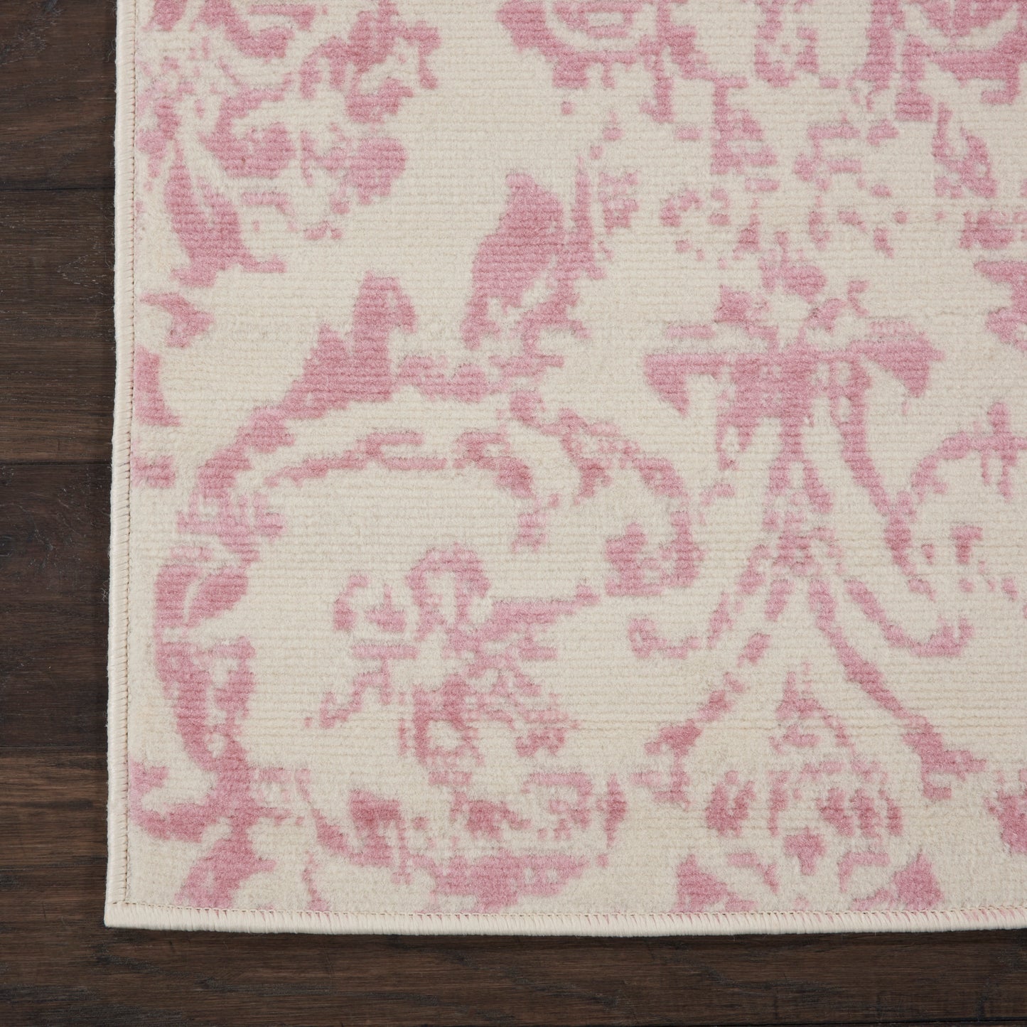 Cream rug with pink damask pattern; woven textile on dark wooden floor, stitched edge, rectangular decorative mat.