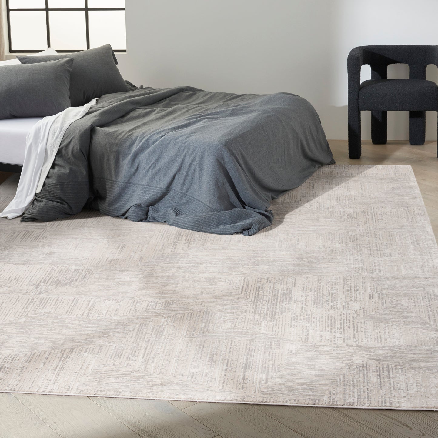 Modern bedroom with dark gray duvet and pillows on a low bed, light textured area rug, and a dark chair.
