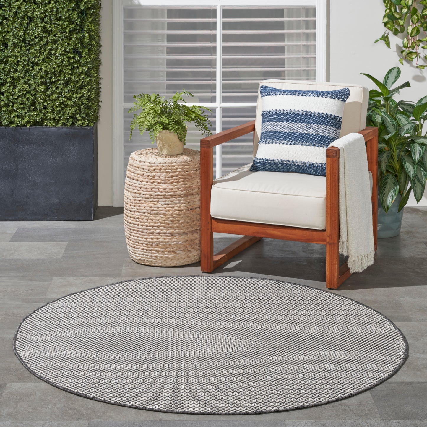 Round gray outdoor rug on a patio; nearby is an outdoor wooden chair with beige cushion and blue striped pillow, plus a wicker plant stand.