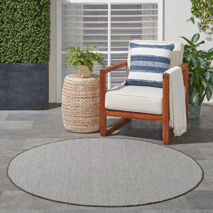 Round gray outdoor rug on a patio; nearby is an outdoor wooden chair with beige cushion and blue striped pillow, plus a wicker plant stand.