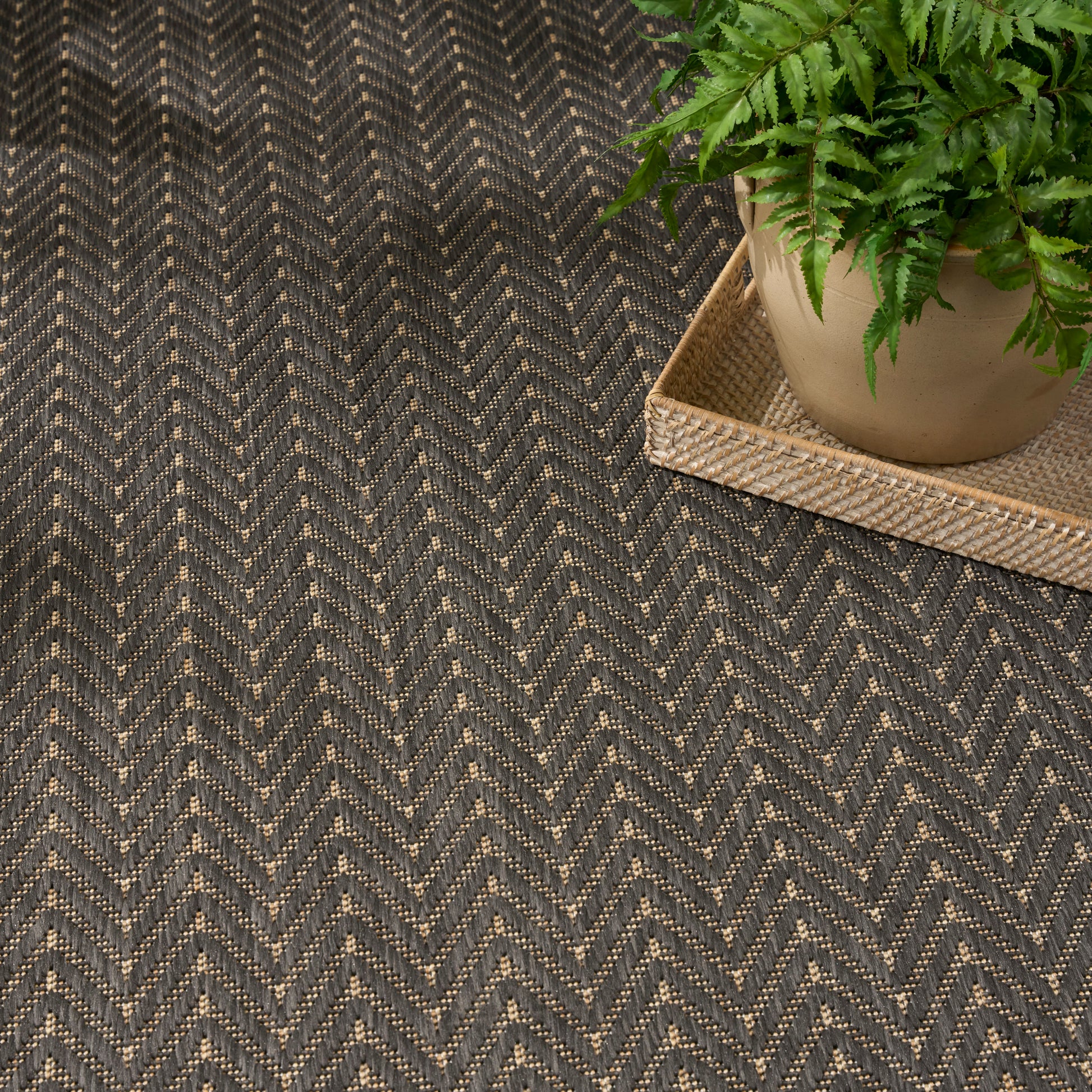 Dark gray & beige chevron woven rug with a potted fern on a natural woven tray.