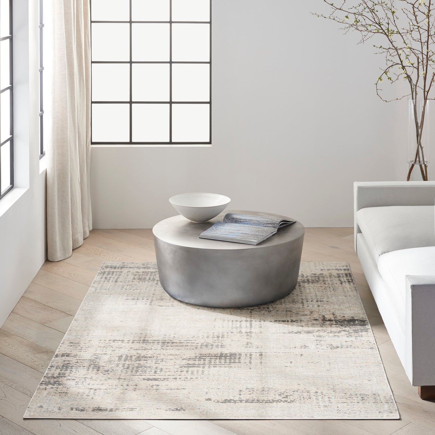 Round gray concrete coffee table with a white bowl & magazine in a bright minimalist living room with a beige rug & grid-window.