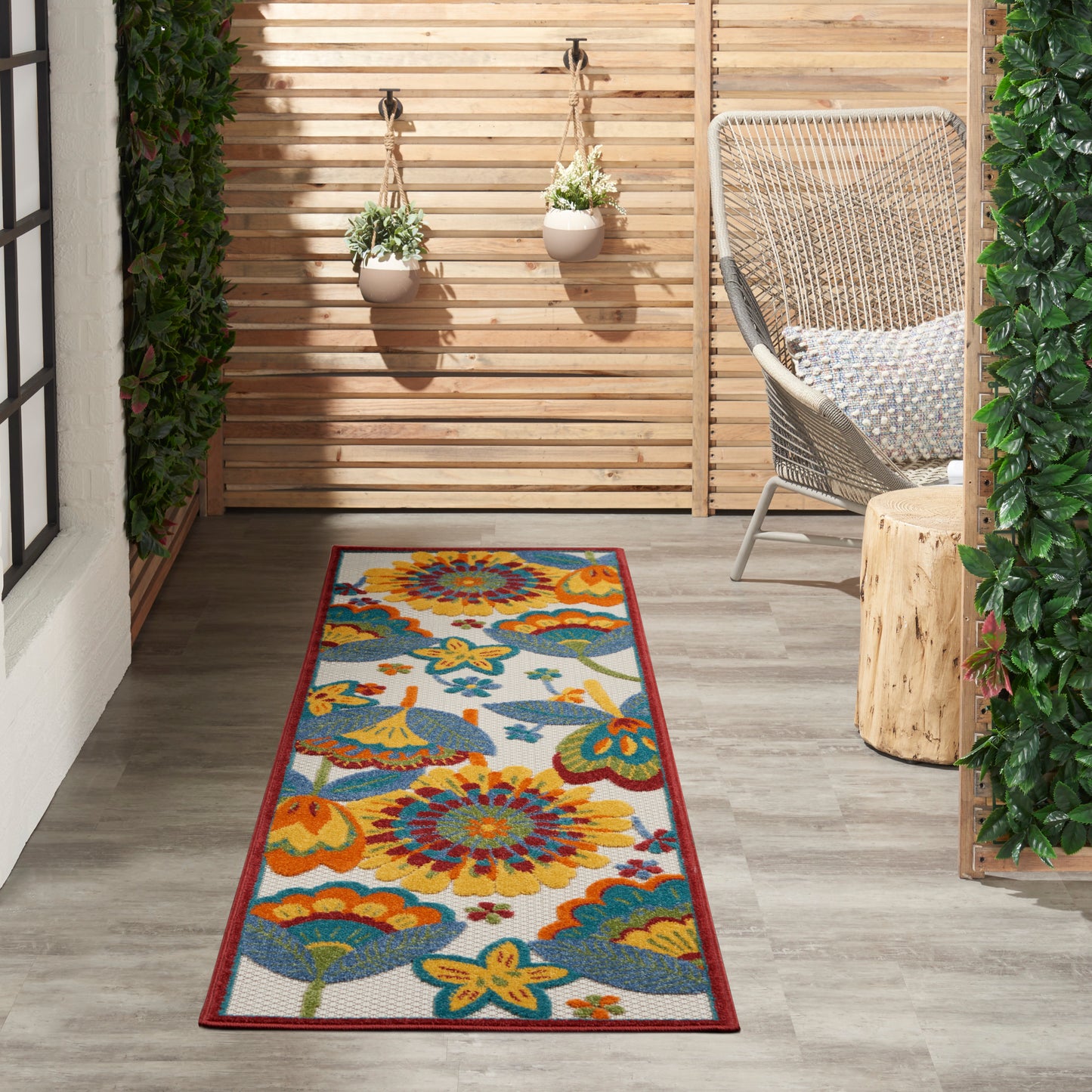 Outdoor runner rug with vibrant yellow, orange, teal flowers and red border on a wooden deck beside a wicker chair.