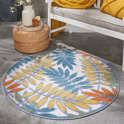 Round rug with blue, yellow, orange tropical leaf pattern on light gray background; indoor living room setting.