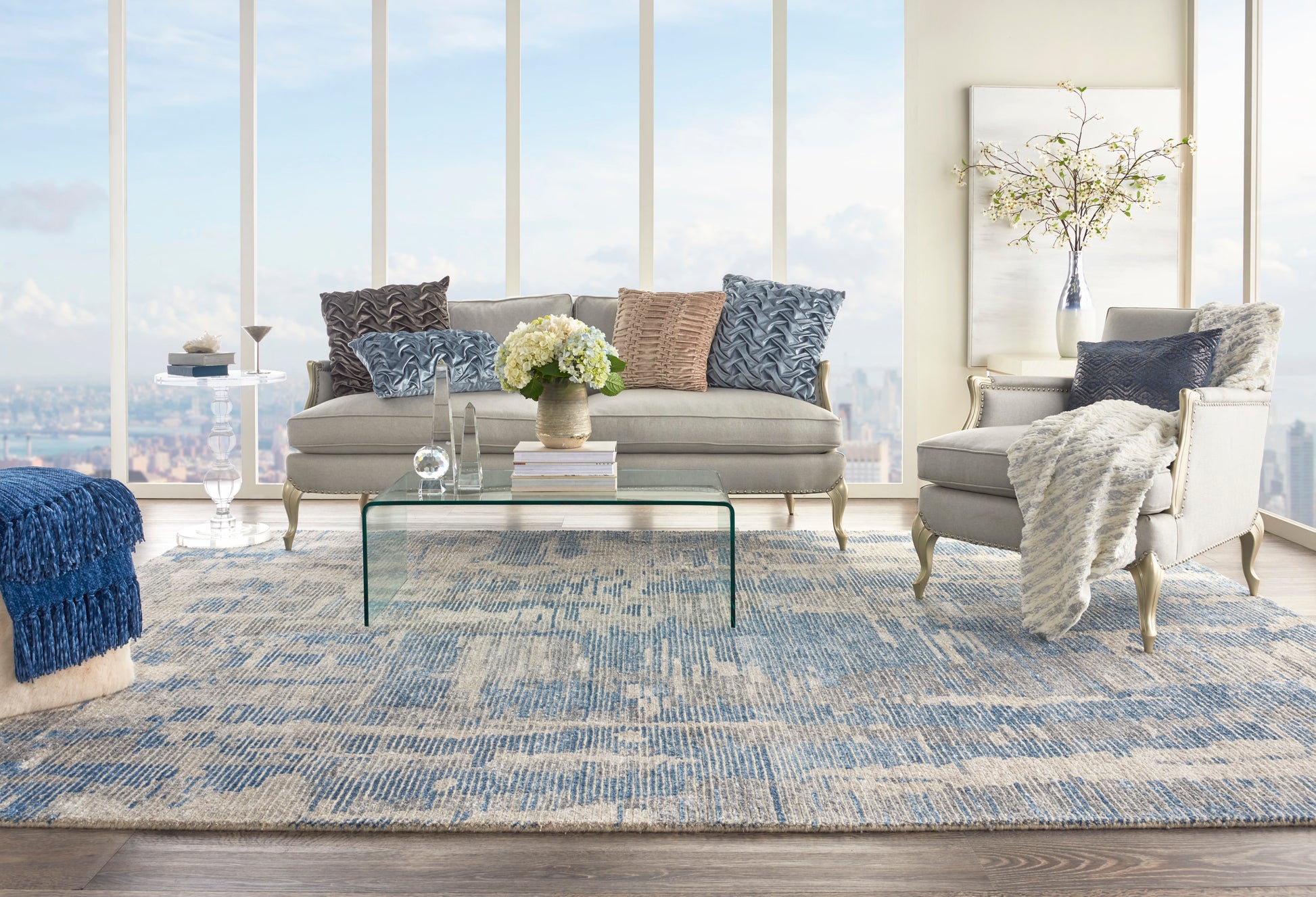 Bright coastal living room with large windows, beige sofa, gray armchair, blue patterned rug, glass coffee table, blue pillows & knit throw.