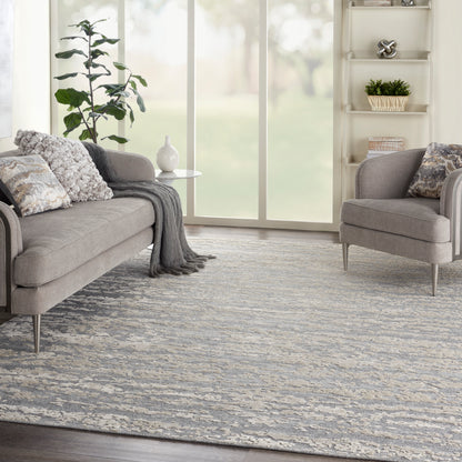 Modern gray living room with a textured blue-gray rug, matching sofa and armchair, throw blanket, plant, and shelves.
