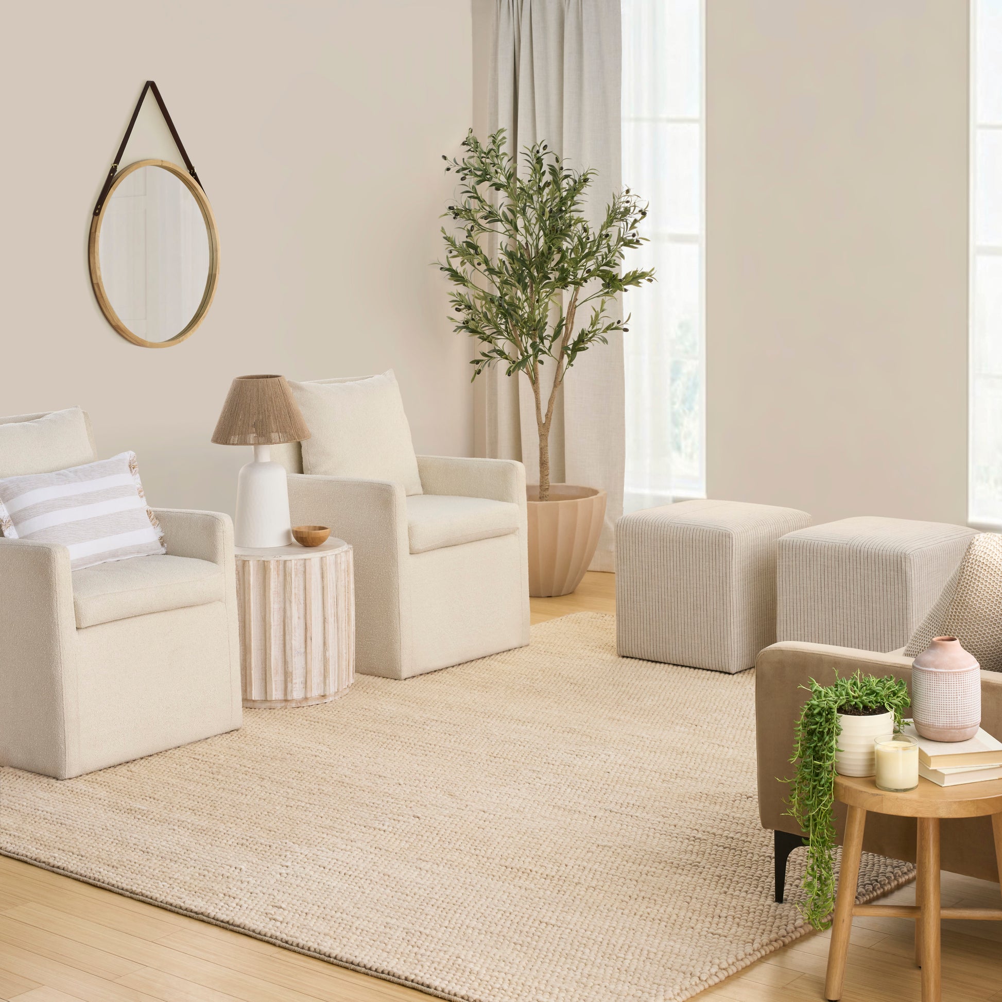 Cream boucle armchairs, round wood side table, woven rug, wall mirror, potted tree and ottomans in a sunlit neutral living room.