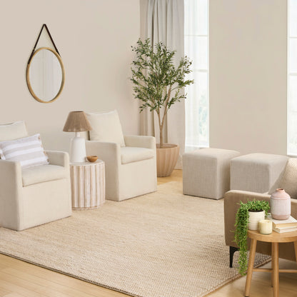 Cream boucle armchairs, round wood side table, woven rug, wall mirror, potted tree and ottomans in a sunlit neutral living room.