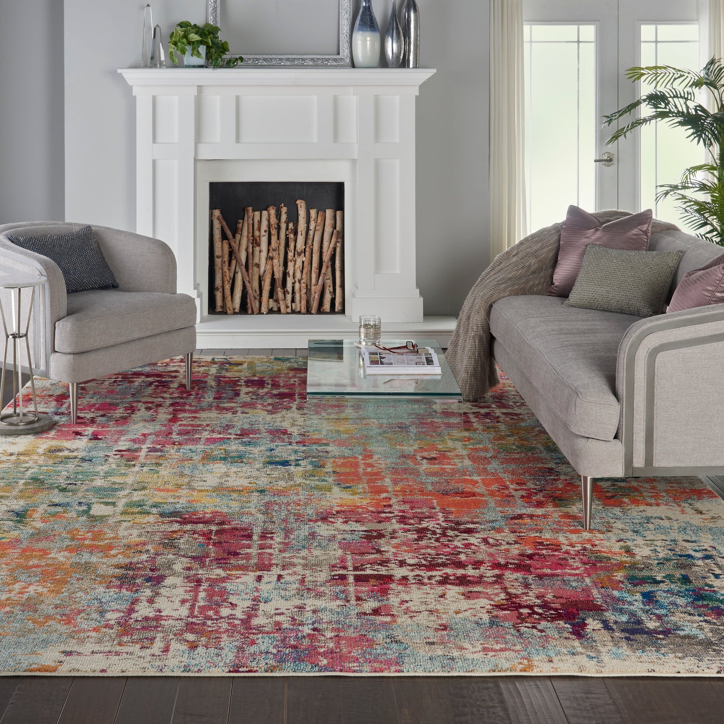 White mantel fireplace with stacked firewood, gray upholstered seating, glass coffee table, & bold multicolor abstract rug.