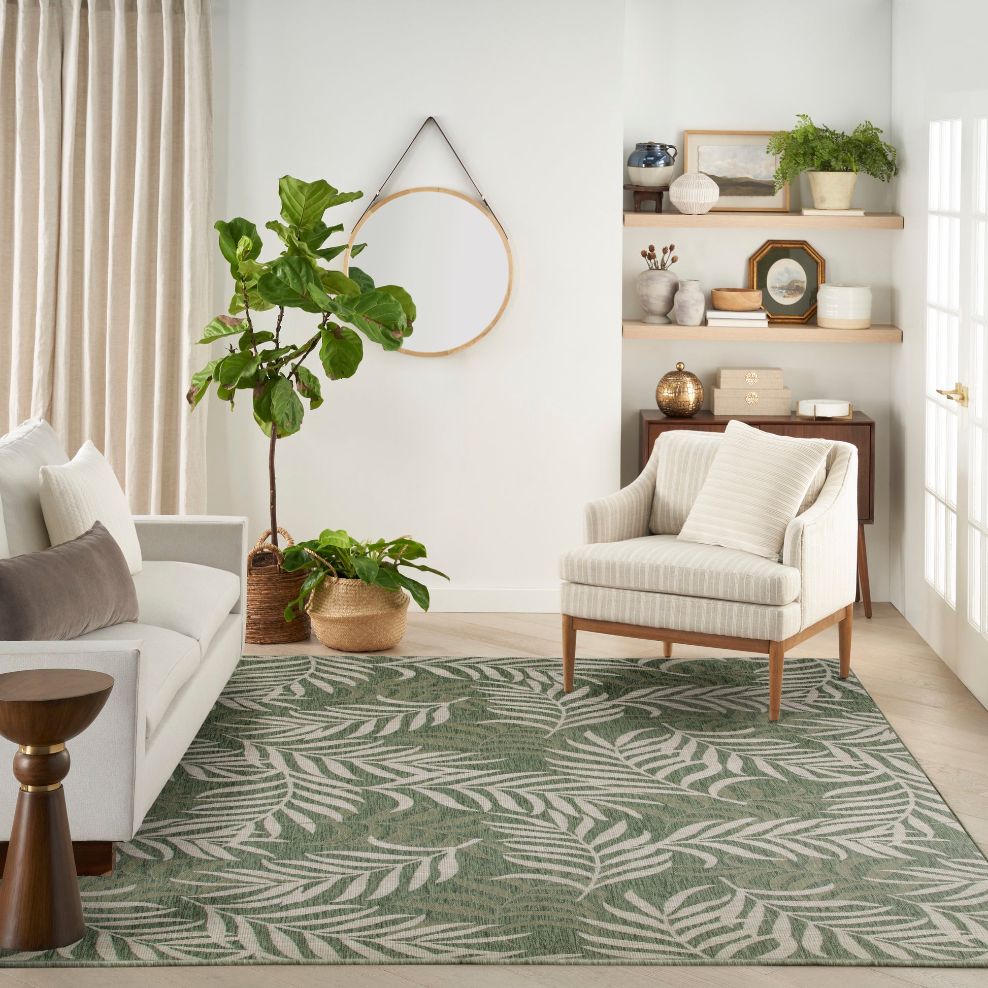 Living room with round wall mirror, green palm-pattern rug, cream sofa, striped armchair, potted plants, beige curtains.