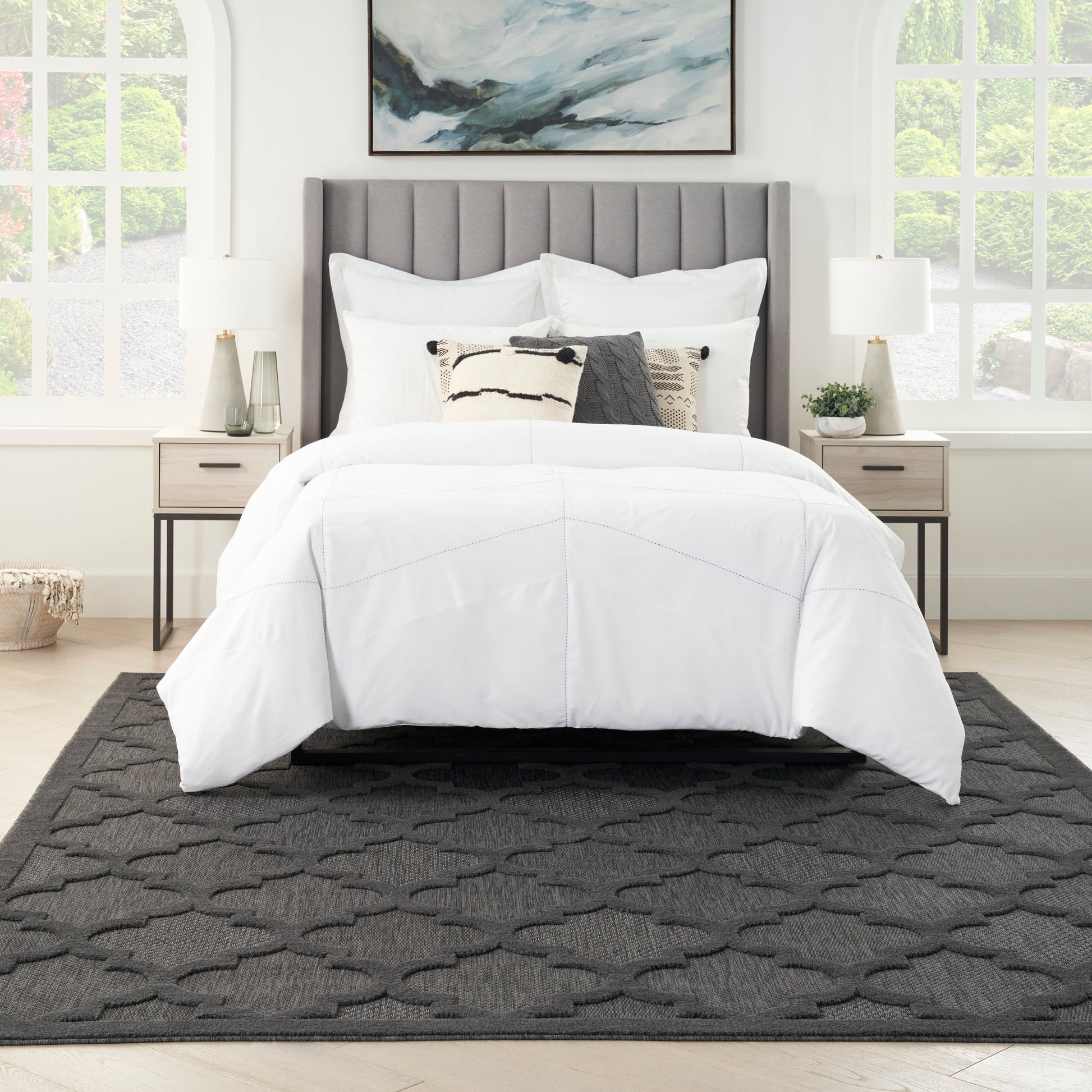 Queen bed with gray tufted headboard and white bedding, lamps on nightstands, abstract art, large windows, dark gray rug.