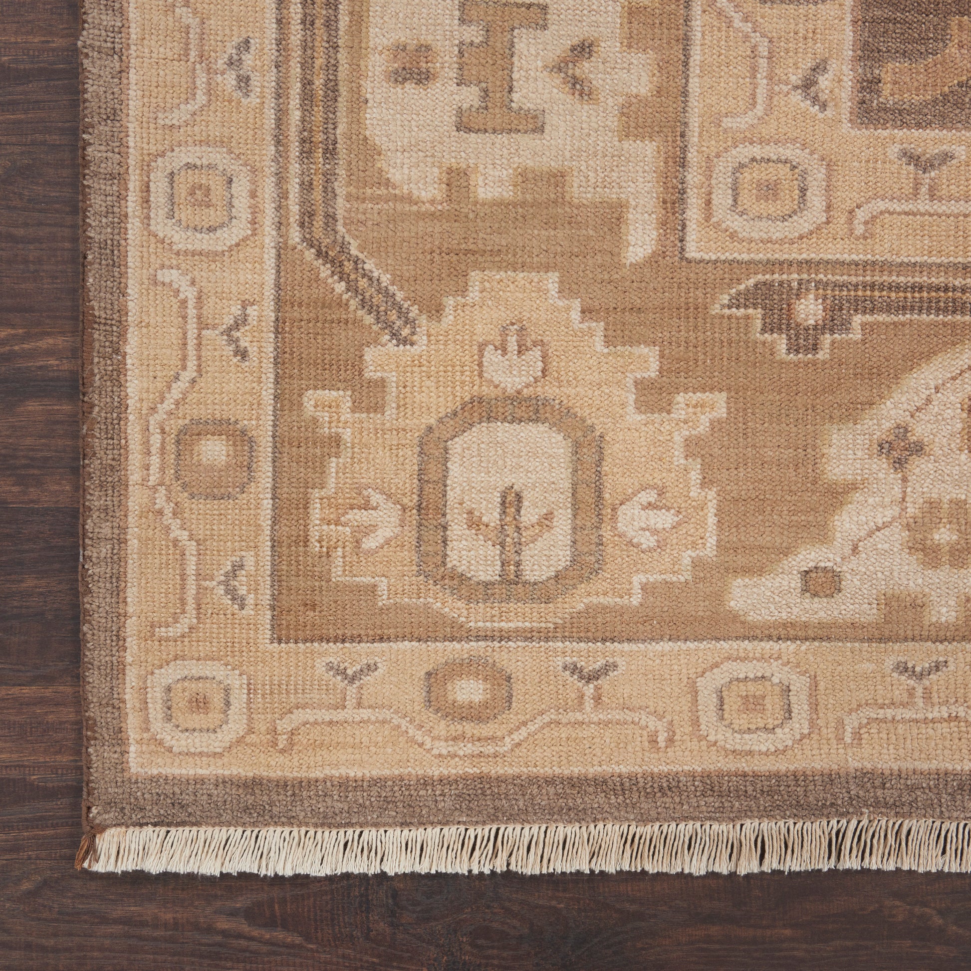 Beige and tan oriental rug with geometric borders and fringe along the edge on dark wood floor.