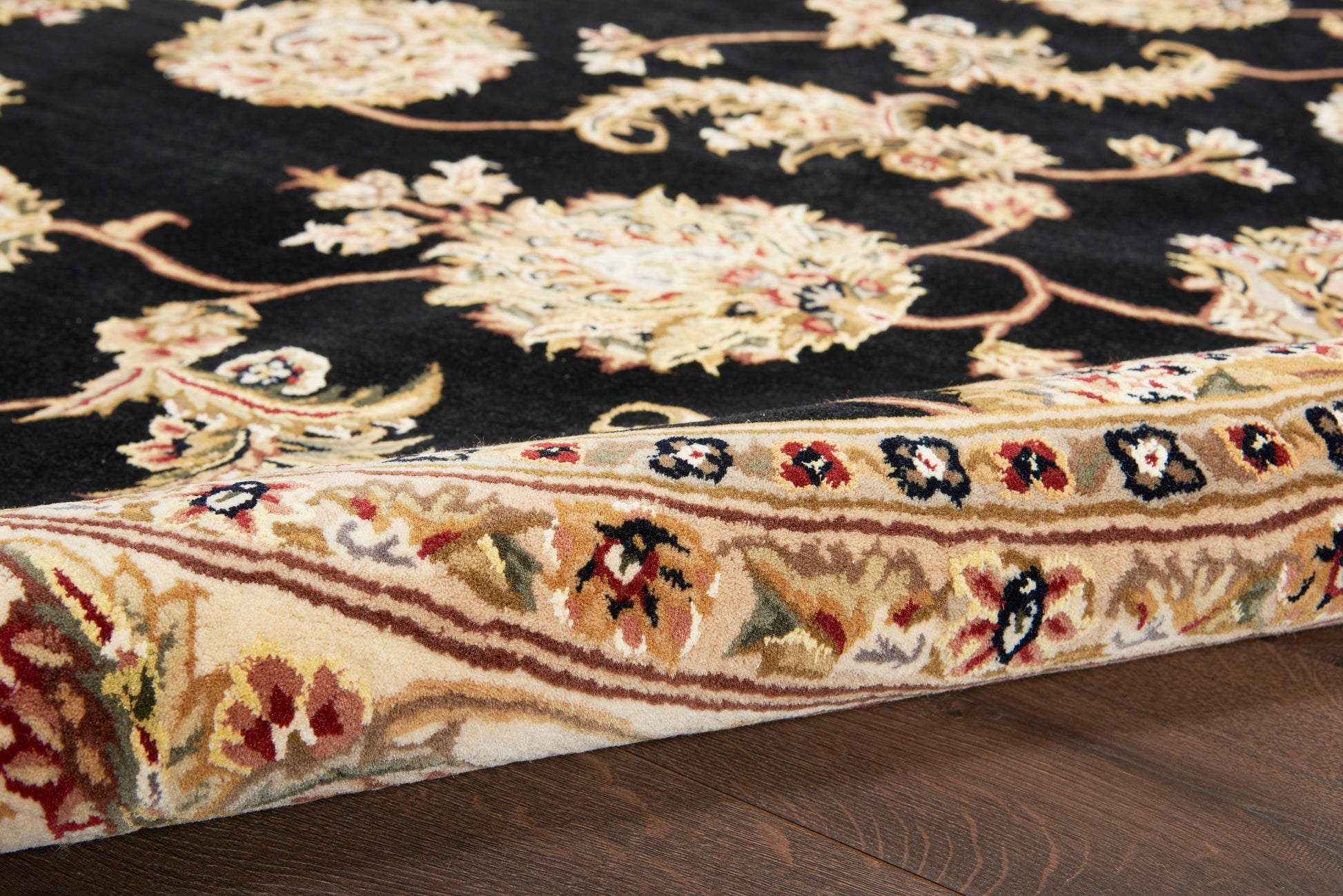 Black oriental rug with ivory, red, and gold floral motifs; thick beige border and rolled edge on dark wood floor.