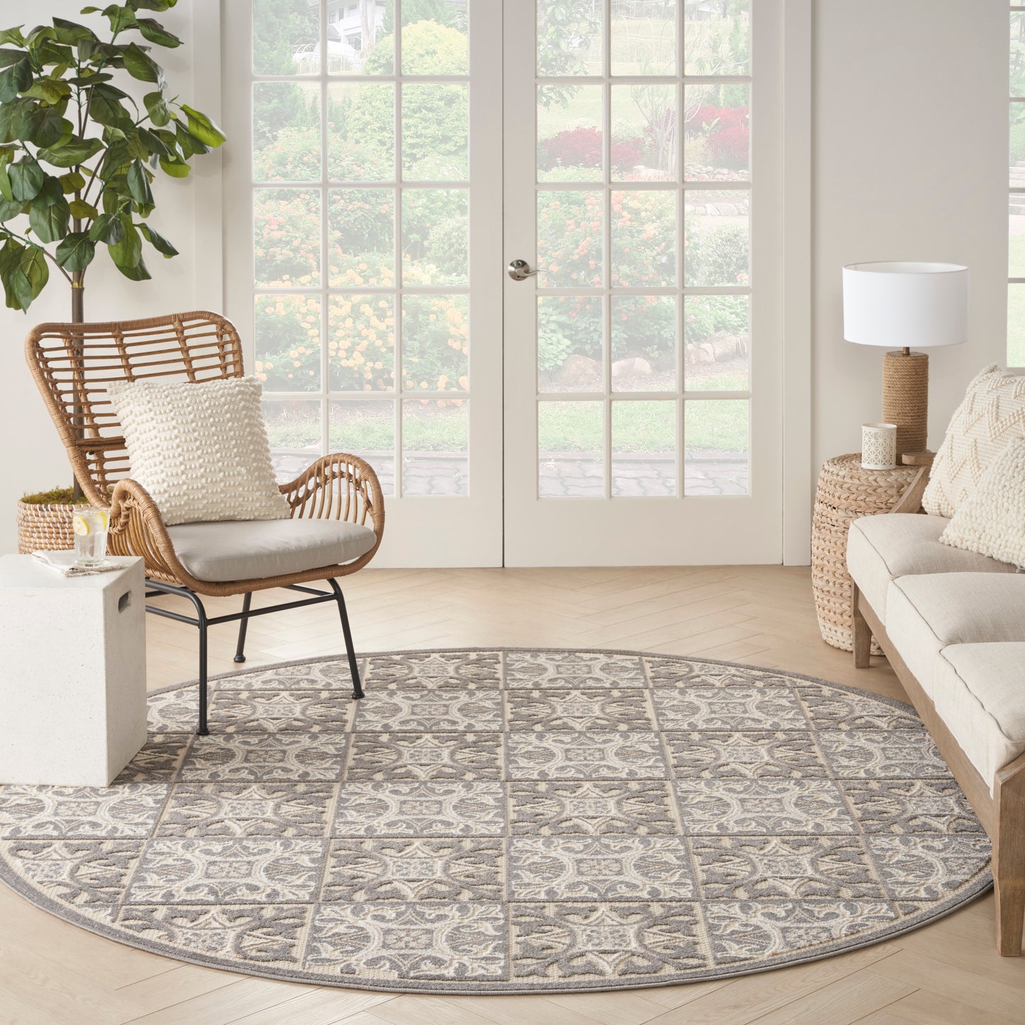 Round gray and taupe patterned area rug on light wood floor, with a woven rattan chair, cream cushions, wicker side table, and sofa.