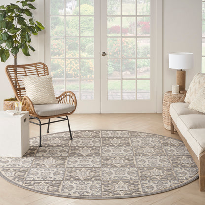 Round gray and taupe patterned area rug on light wood floor, with a woven rattan chair, cream cushions, wicker side table, and sofa.