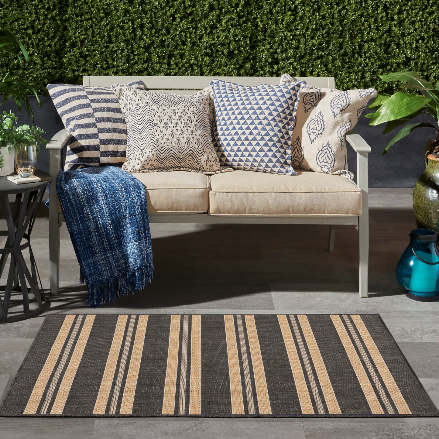 Outdoor beige rug (3' x 5') with a striped pattern, greenery backdrop and lantern accents.