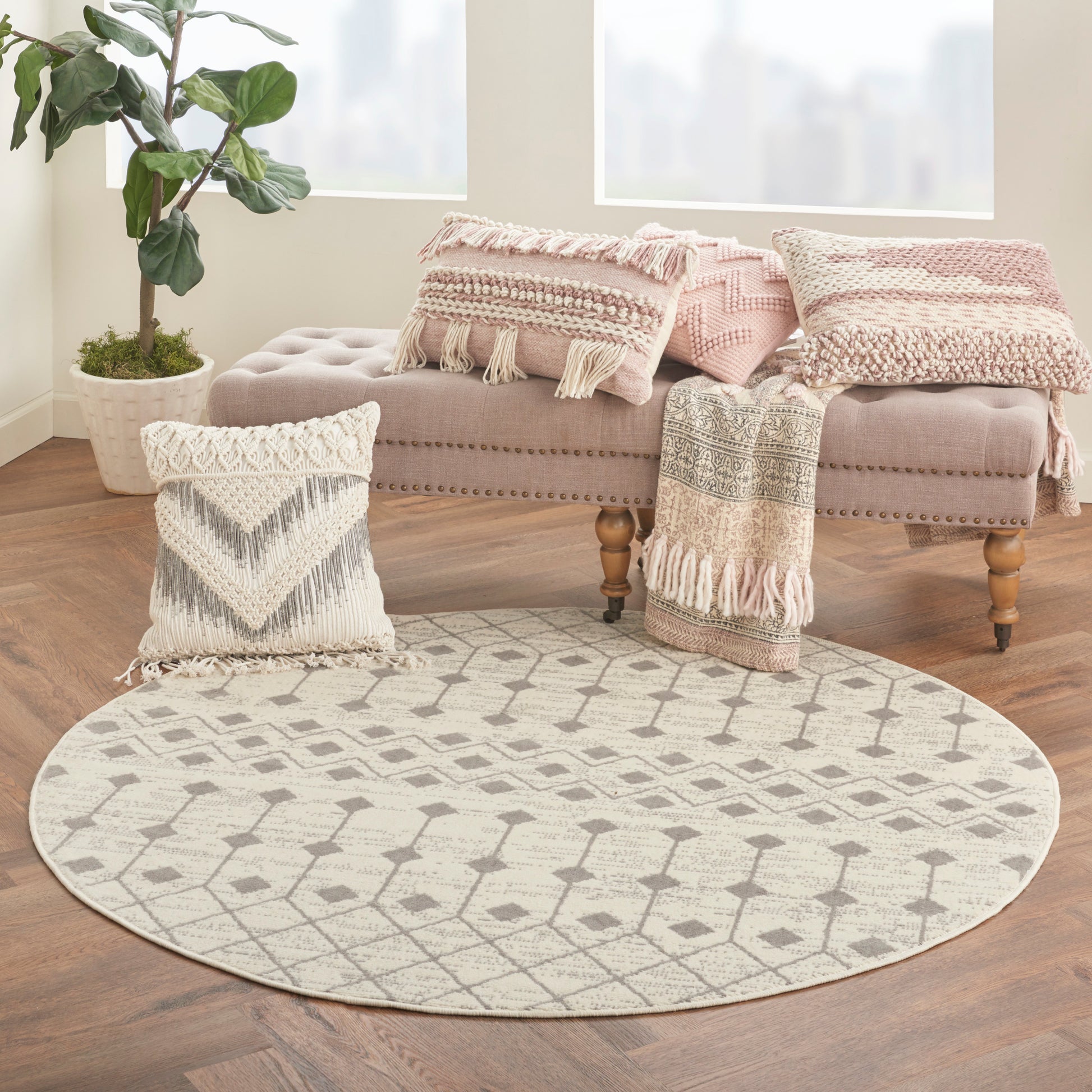 Round beige geometric rug on a wooden floor with taupe tufted bench, pink woven throws, chevron pillow, and a potted plant.