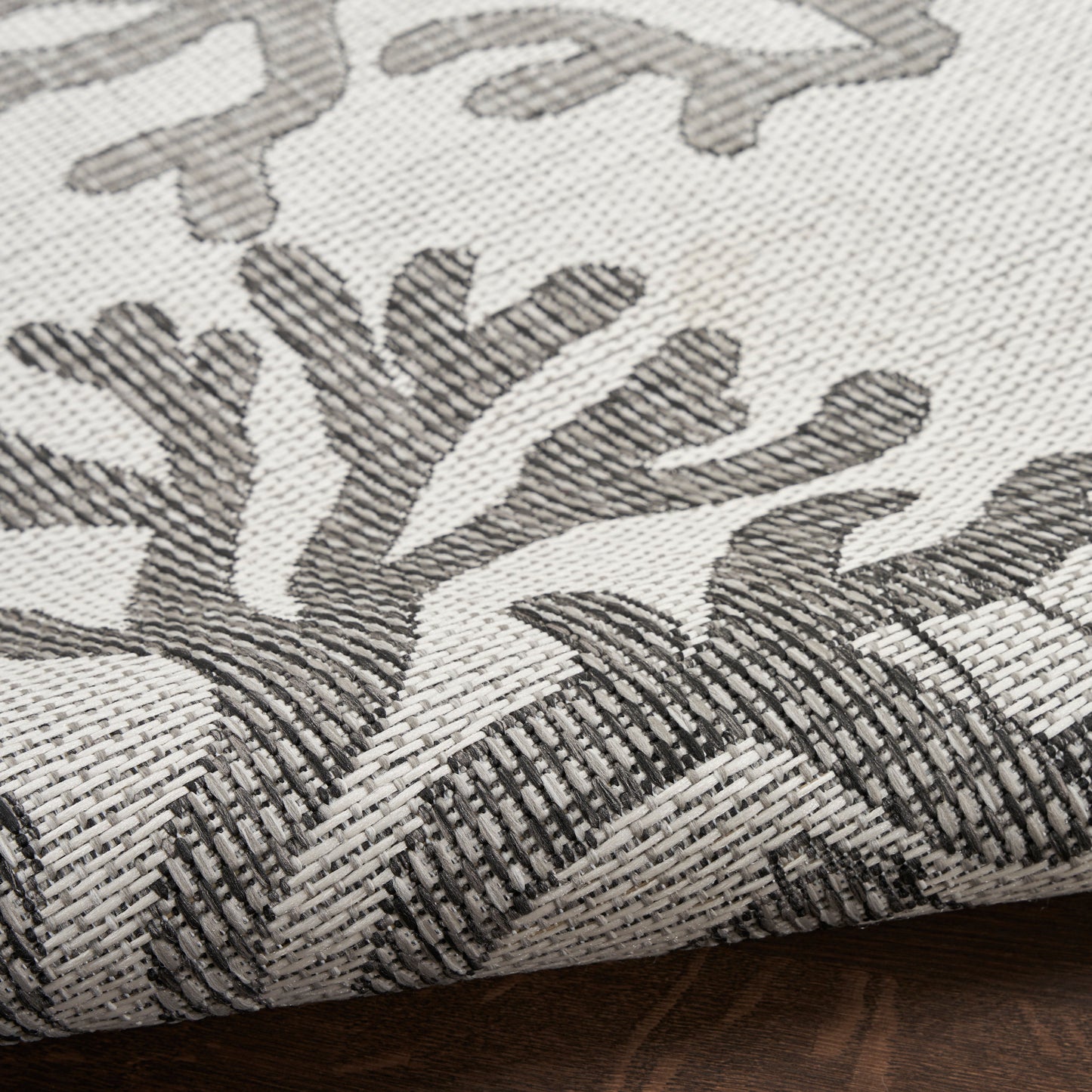 Beige woven rug with dark gray botanical leaf pattern and a rolled edge; textured diagonal weave.