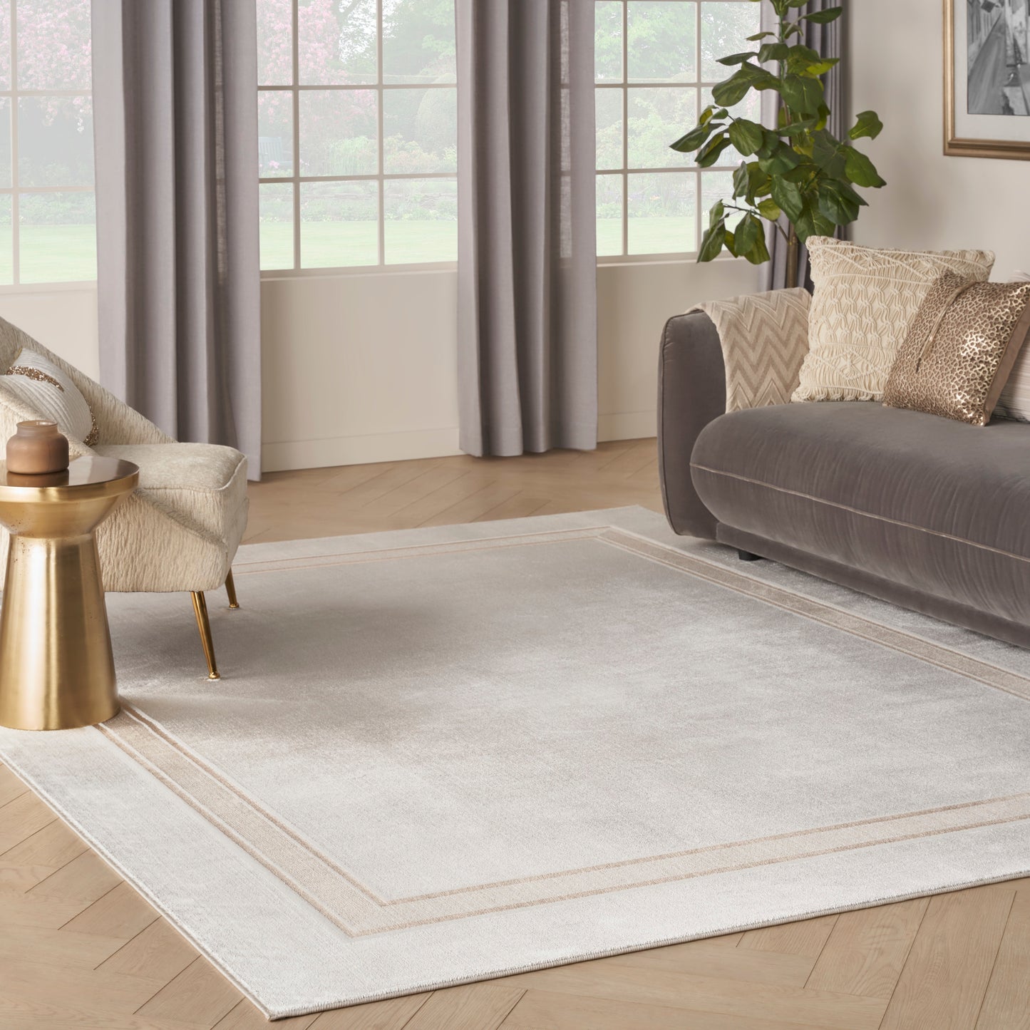 Light gray area rug with beige border in a modern living room featuring a gray sofa, gold side table, and large windows.