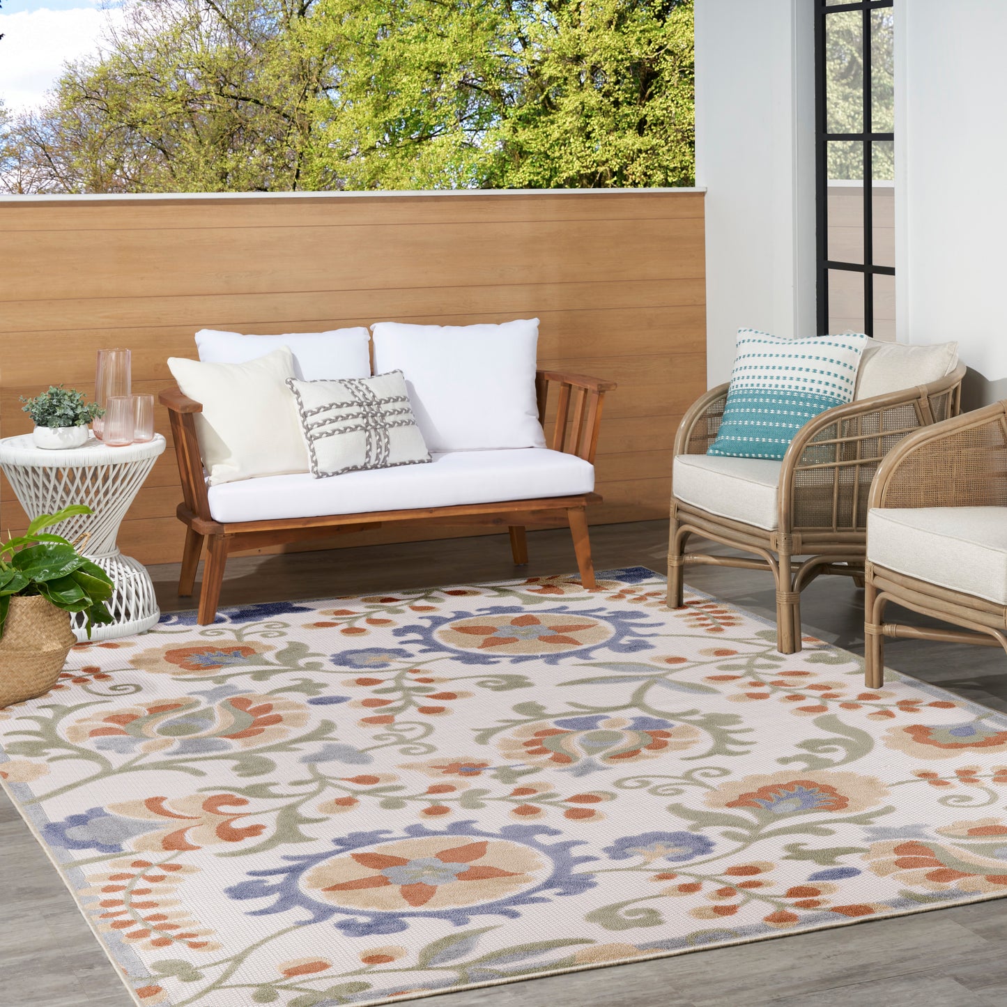 Outdoor patio with a wood-framed sofa, wicker chairs, white cushions, and a colorful floral rug; potted plant and glass door.