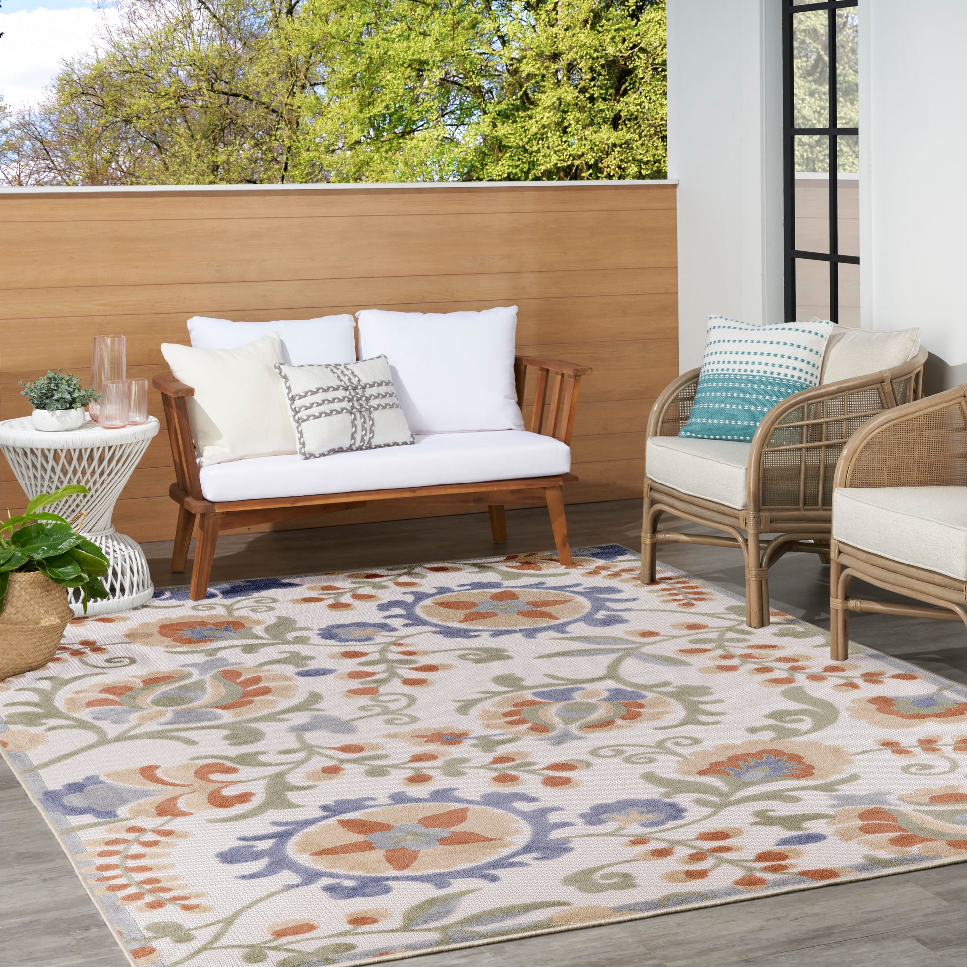 Outdoor patio with a wood-framed sofa, wicker chairs, white cushions, and a colorful floral rug; potted plant and glass door.