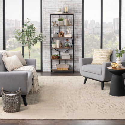 Modern living room with gray armchairs, beige geometric rug, and a tall black metal bookshelf with plants against a brick wall.