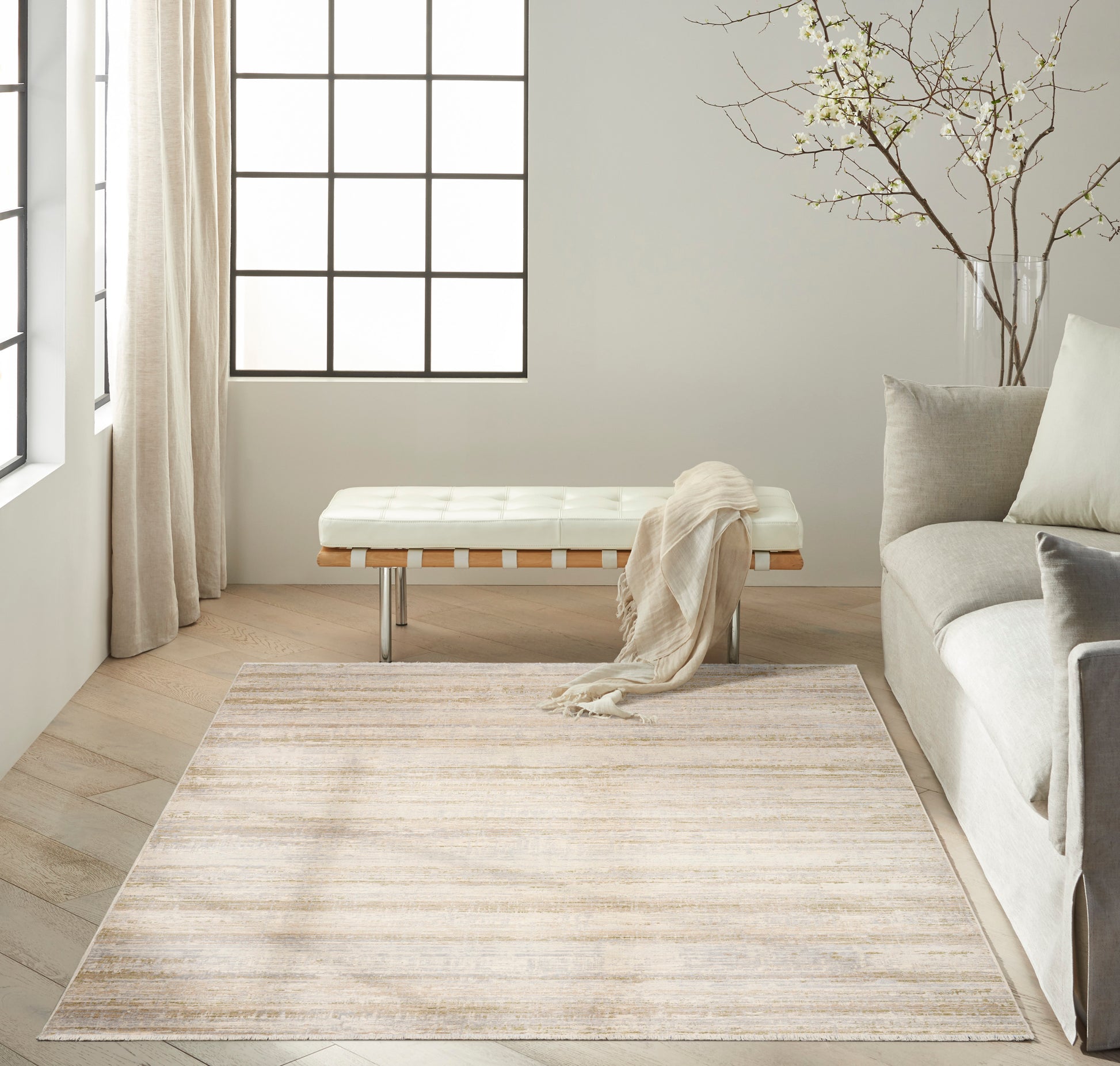 Neutral living room with cream tufted bench, beige rug, light wood floor, gray sofa, sheer curtains, & a vase of branches.