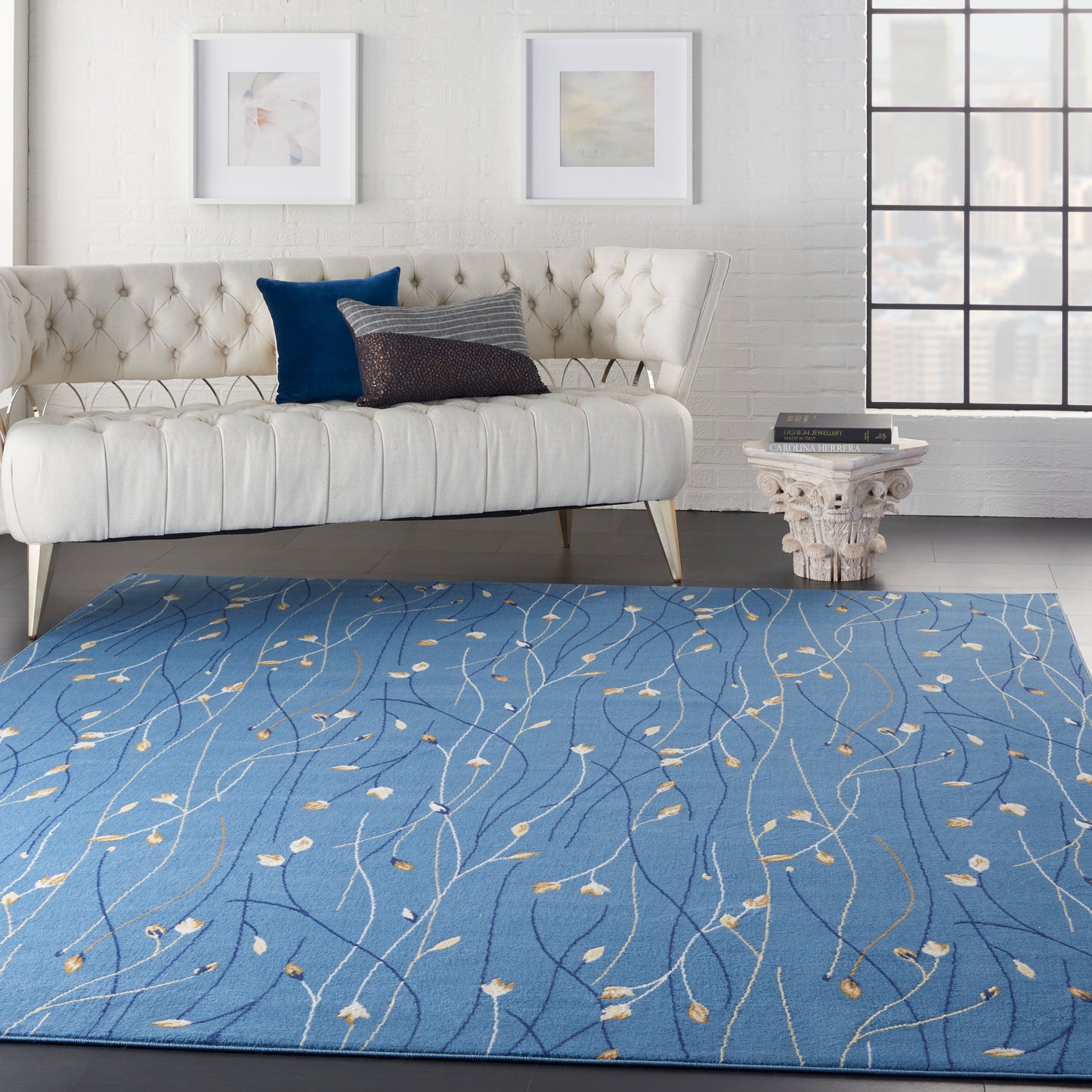 Blue area rug with light beige leaf-vine pattern in a modern living room; white tufted sofa and ornate pedestal side table.