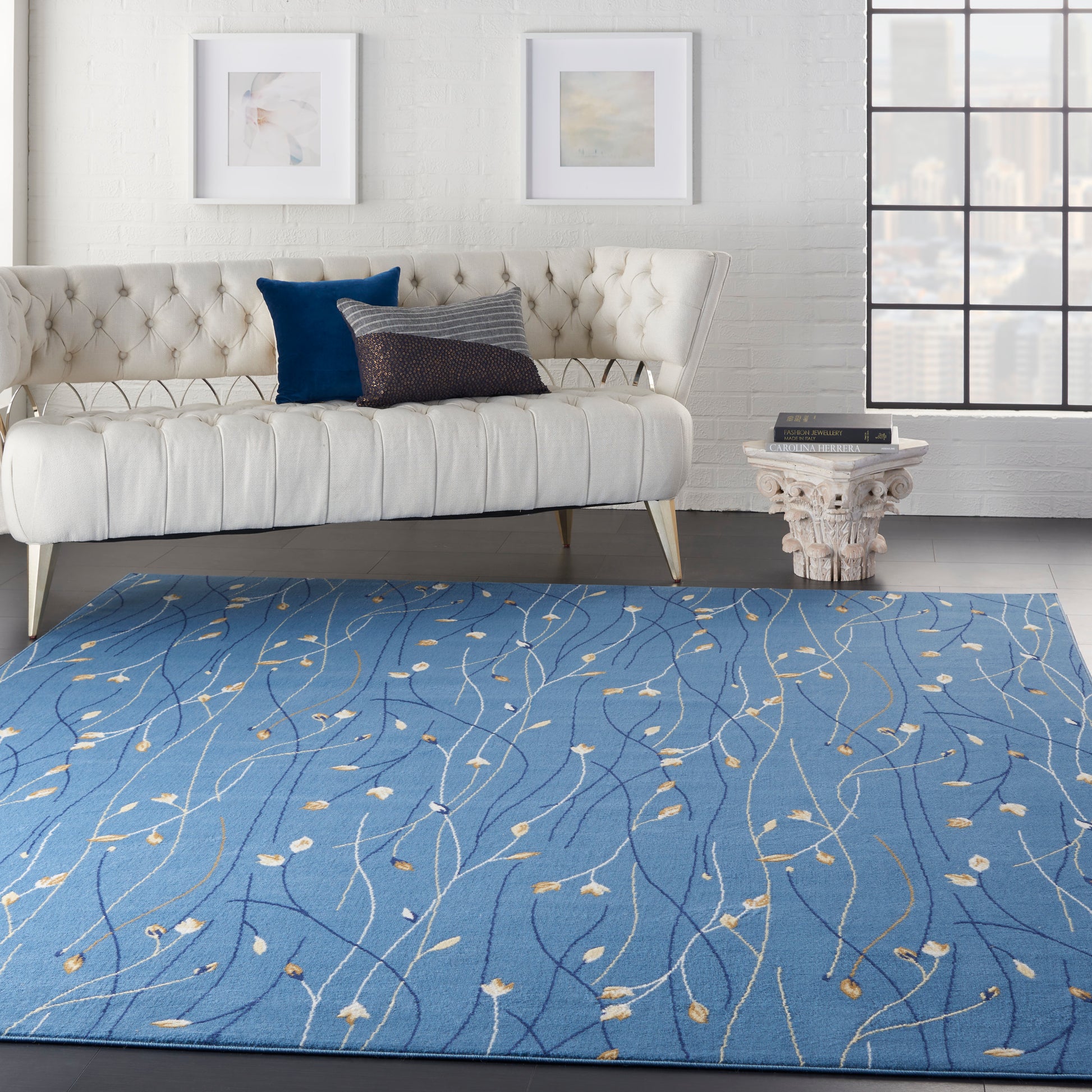 Blue area rug with light beige leaf-vine pattern in a modern living room; white tufted sofa and ornate pedestal side table.