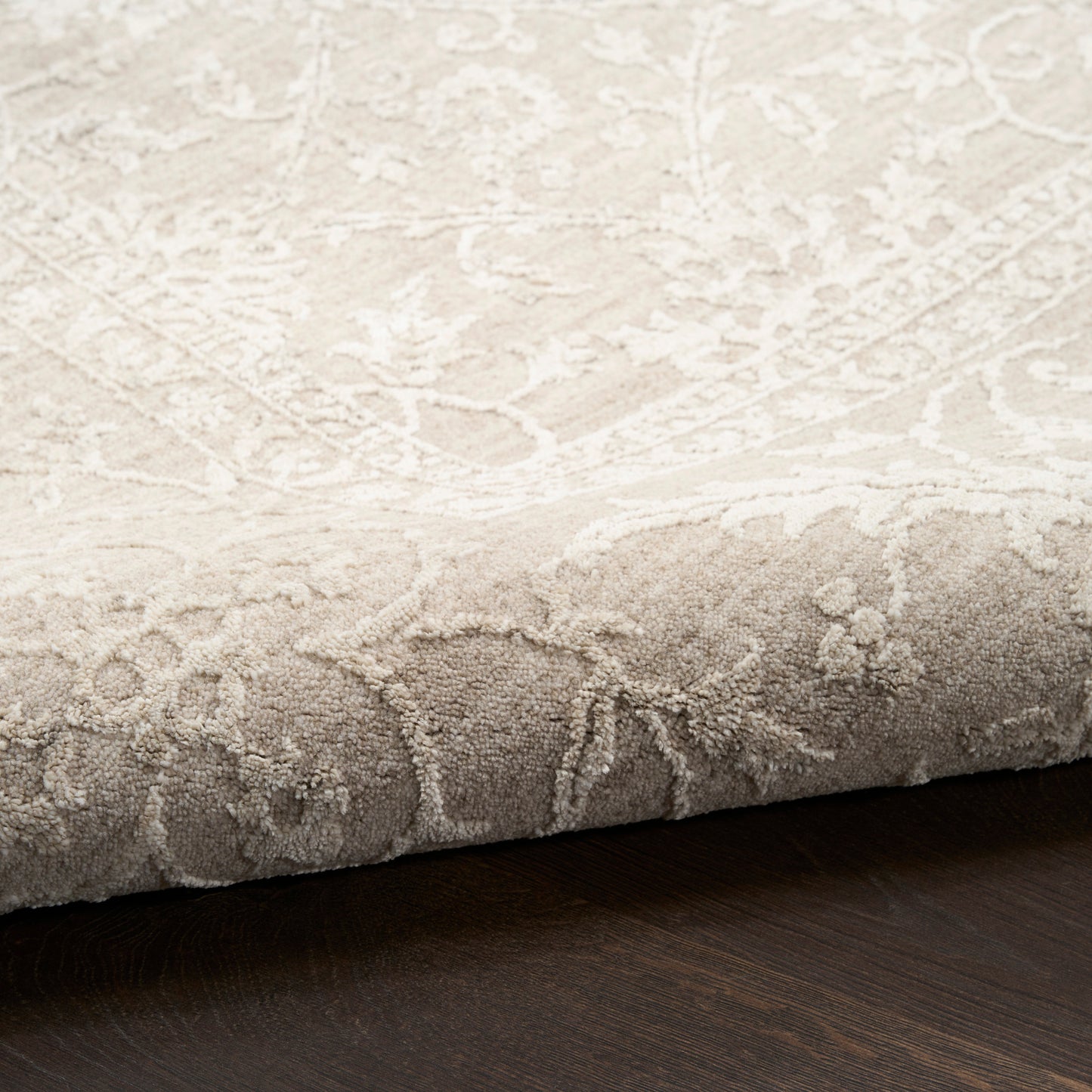 Beige damask patterned area rug with soft plush pile and ivory floral motif, folded edge on dark wood floor.