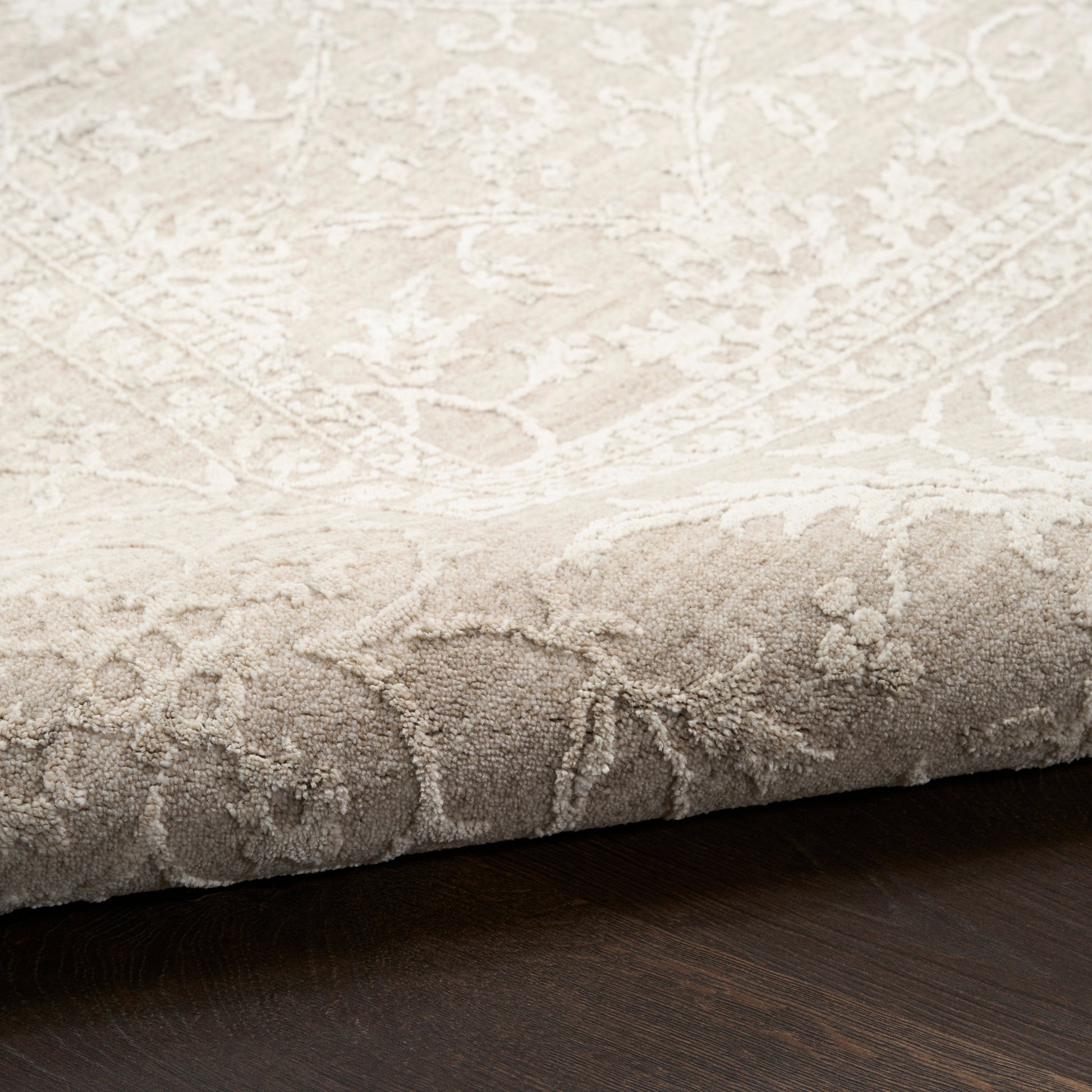 Beige damask patterned area rug with soft plush pile and ivory floral motif, folded edge on dark wood floor.