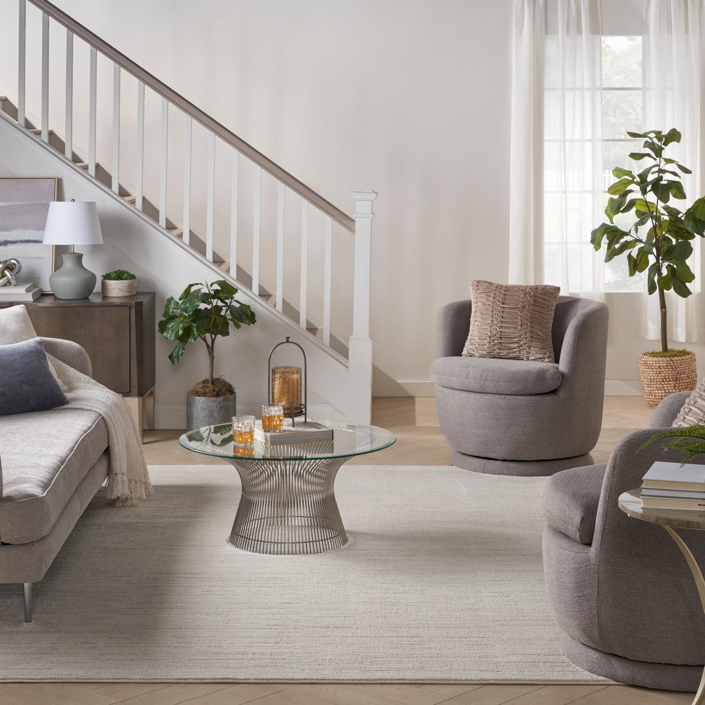 Neutral living room with gray fabric sofa and swivel chairs, round glass coffee table, rug, and greenery.