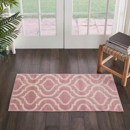 Pink fabric rug with white geometric motif on dark wood floor by glass doors; plant left, stool with magazines right.